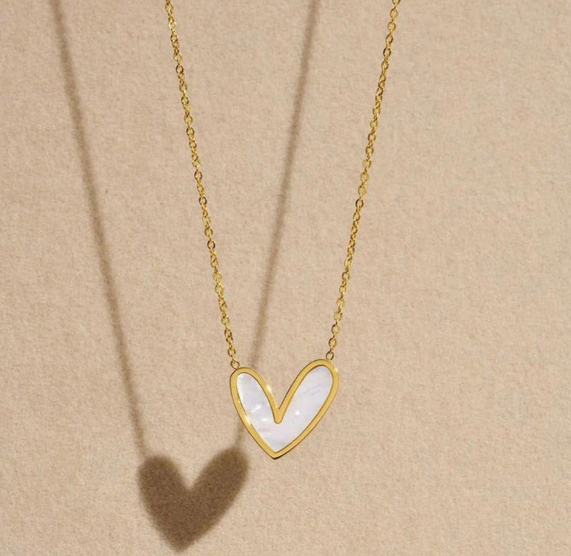ELL DESIGNS gold chain necklace with white heart-shaped pendant casting shadow on beige background