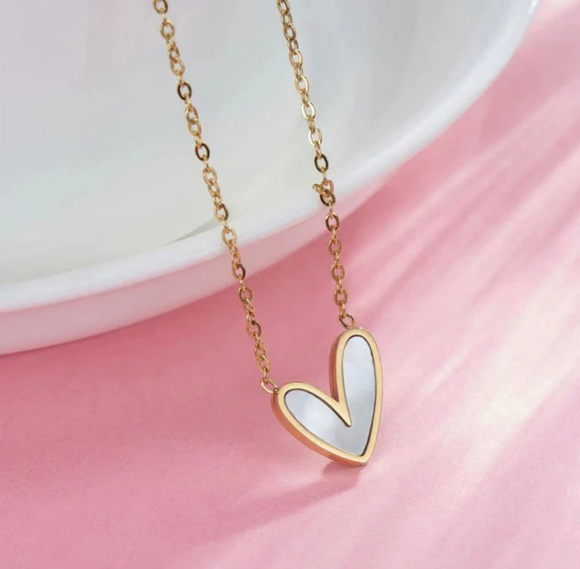 Gold chain necklace with a heart-shaped mother of pearl pendant by ELL DESIGNS on pink background
